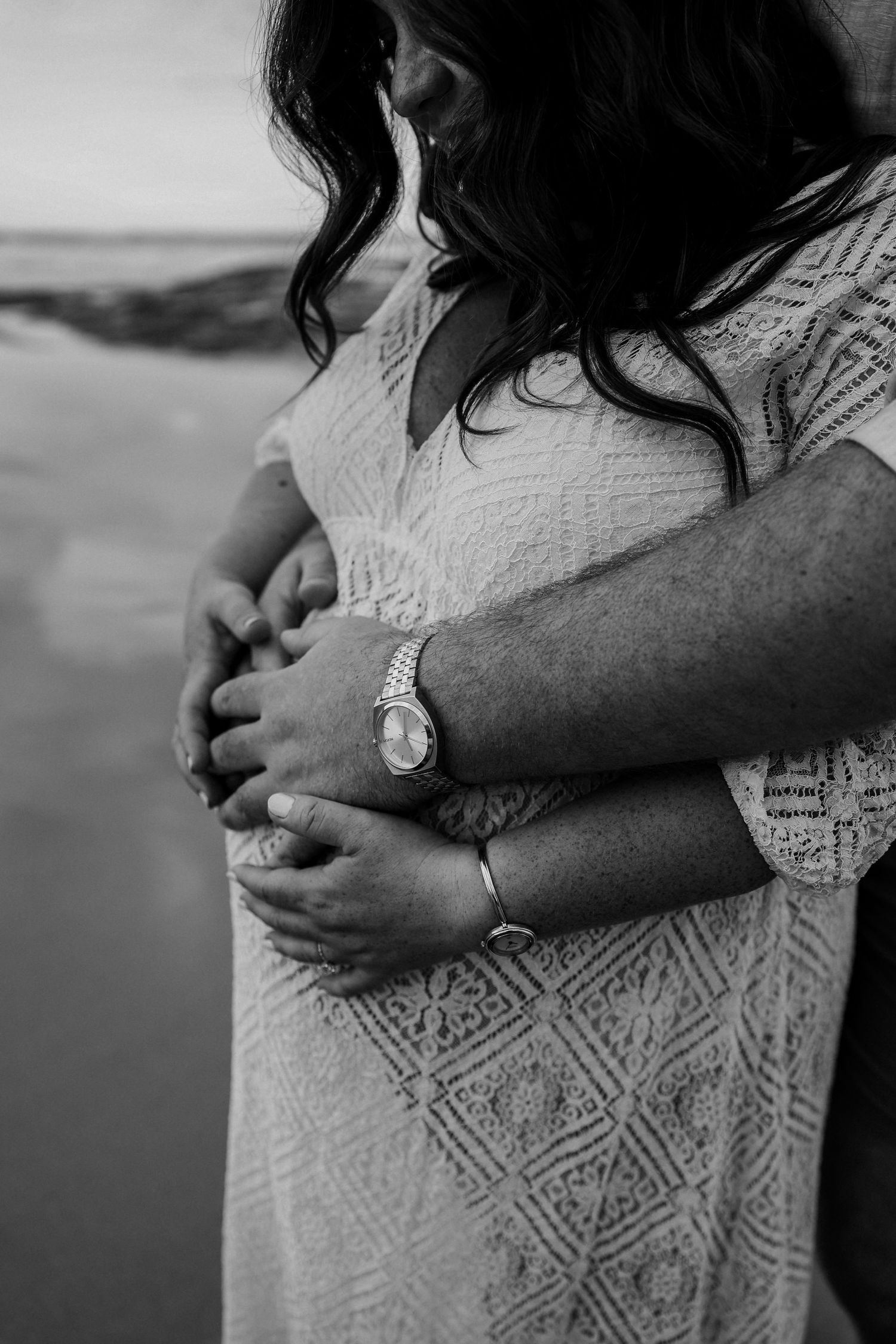Black and white maternity photo of couple embracing pregnant belly on beach, man wearing Rolex watch.