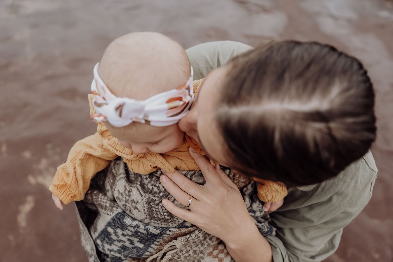 A mother tenderly kisses her newborn baby wrapped in a mustard blanket with a white bow headband outdoors.
