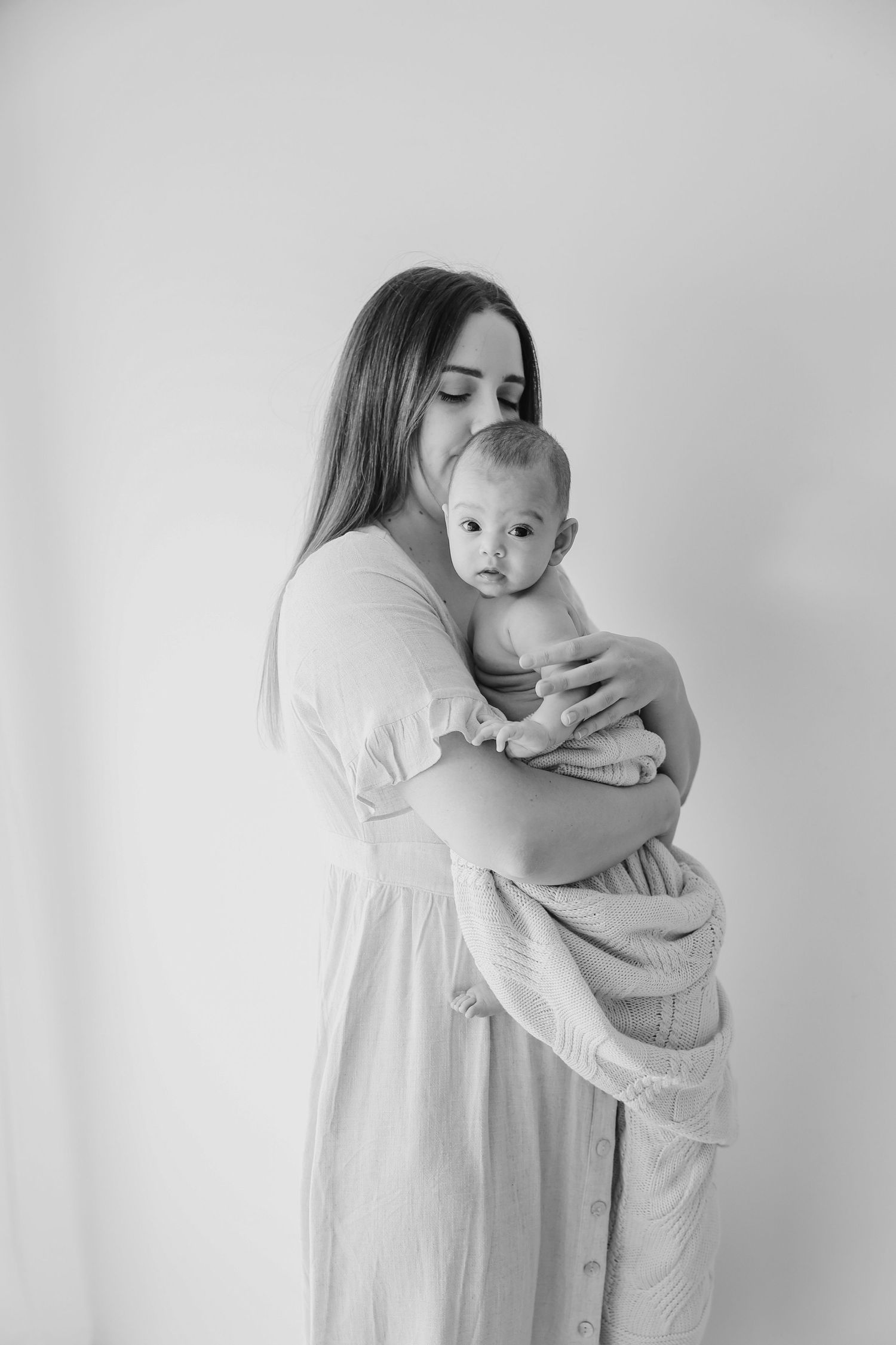 Black and white photo of a mother in a white dress tenderly holding her newborn baby wrapped in a soft blanket.