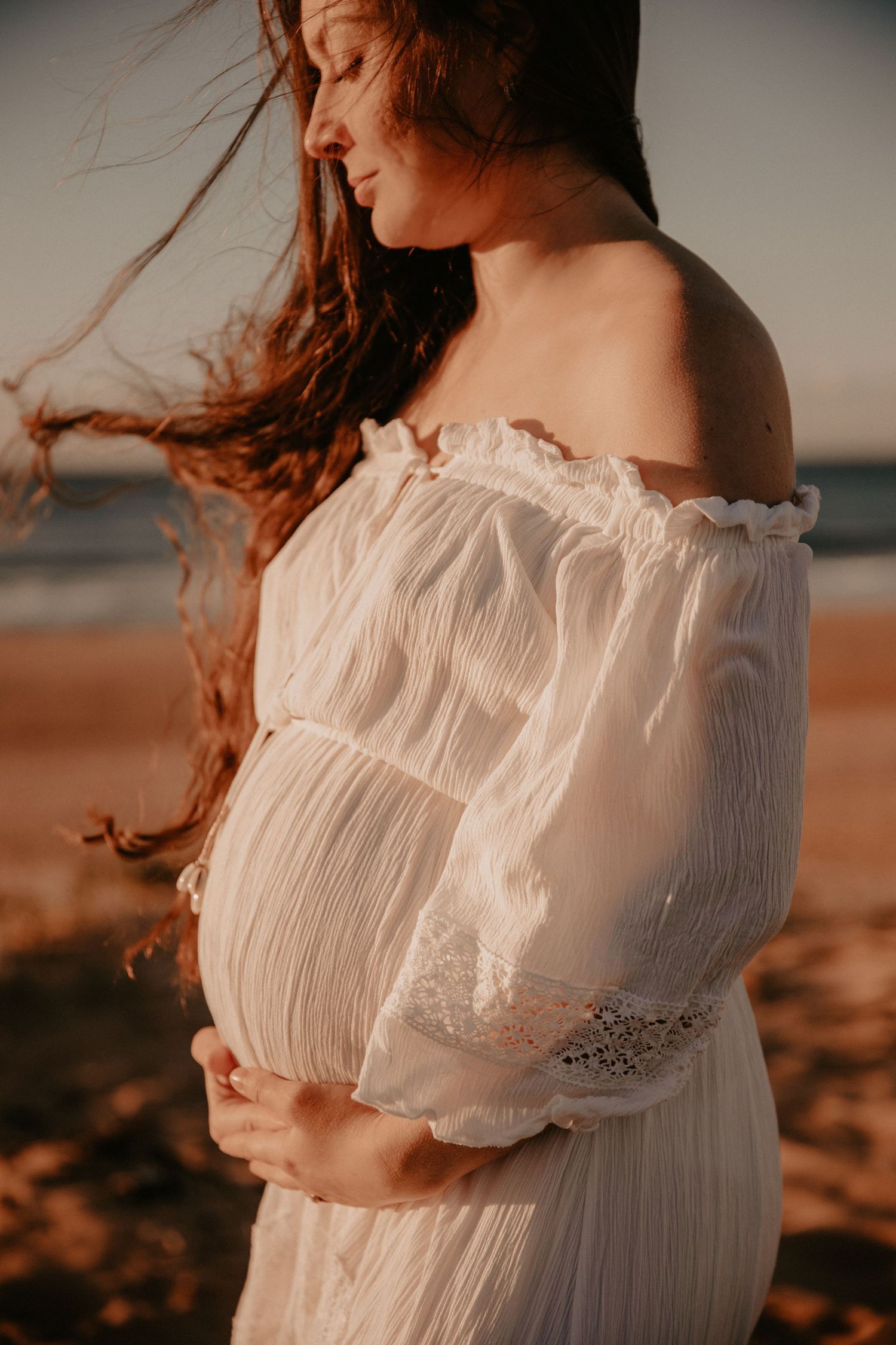 Side profile of pregnant woman in white off-shoulder dress cradling bump on sandy beach.