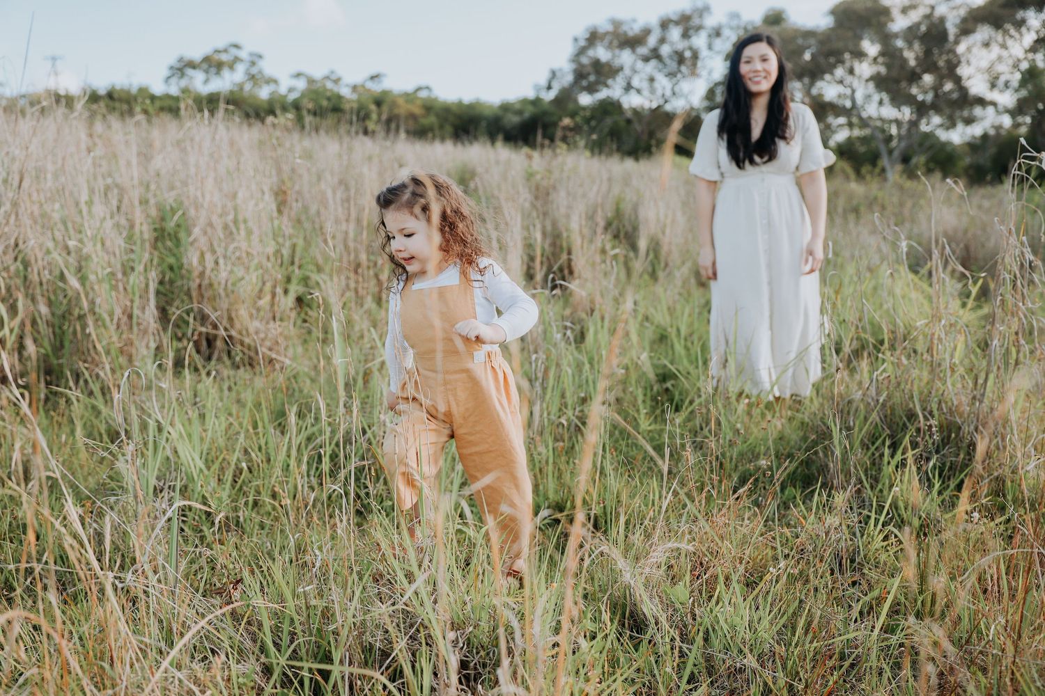 Young girl in yellow overalls explores tall grass field while mother watches nearby.