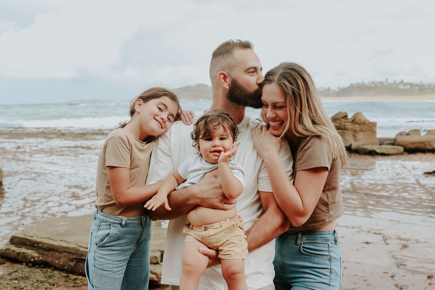 Happy family of four posing on a rocky beach, parents kissing baby while older sister leans in affectionately.