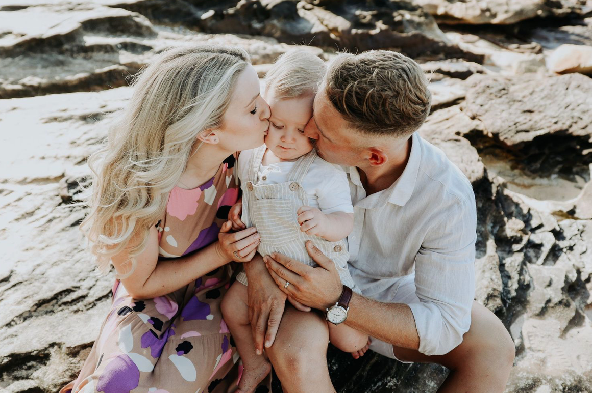 Two parents simultaneously kissing their smiling toddler on the cheeks at a rocky beach.