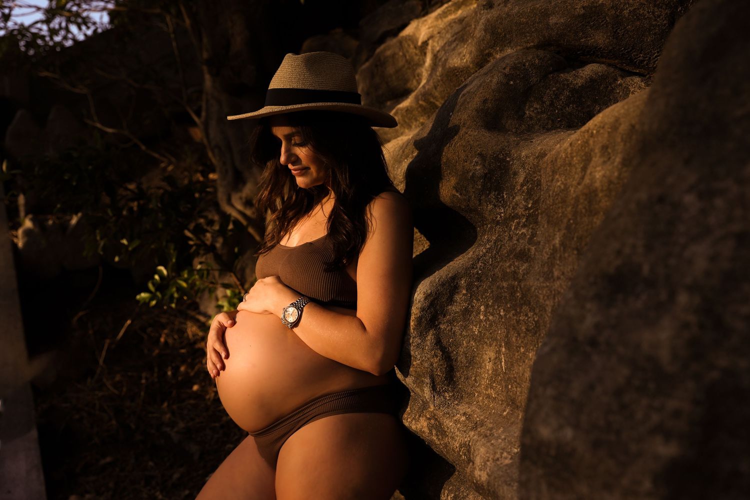 Pregnant woman in brown bikini and tan hat cradling baby bump against rocky backdrop in warm golden light.