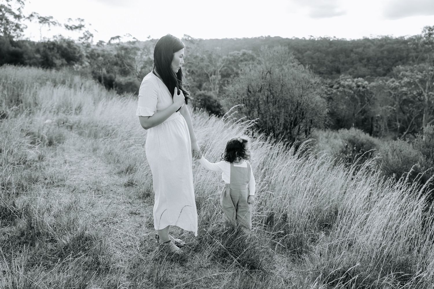 Pregnant woman in white dress holds toddlers hand in grassy field, black and white maternity photo outdoors.