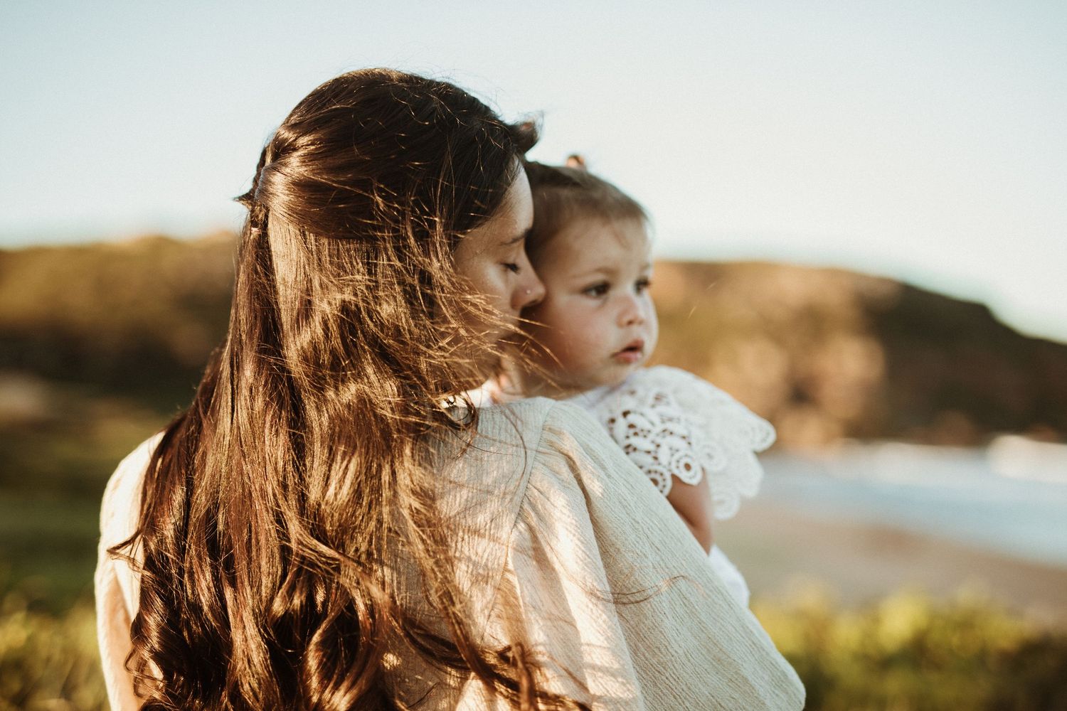 Mother with long dark hair tenderly holds baby girl near coastal cliffs.