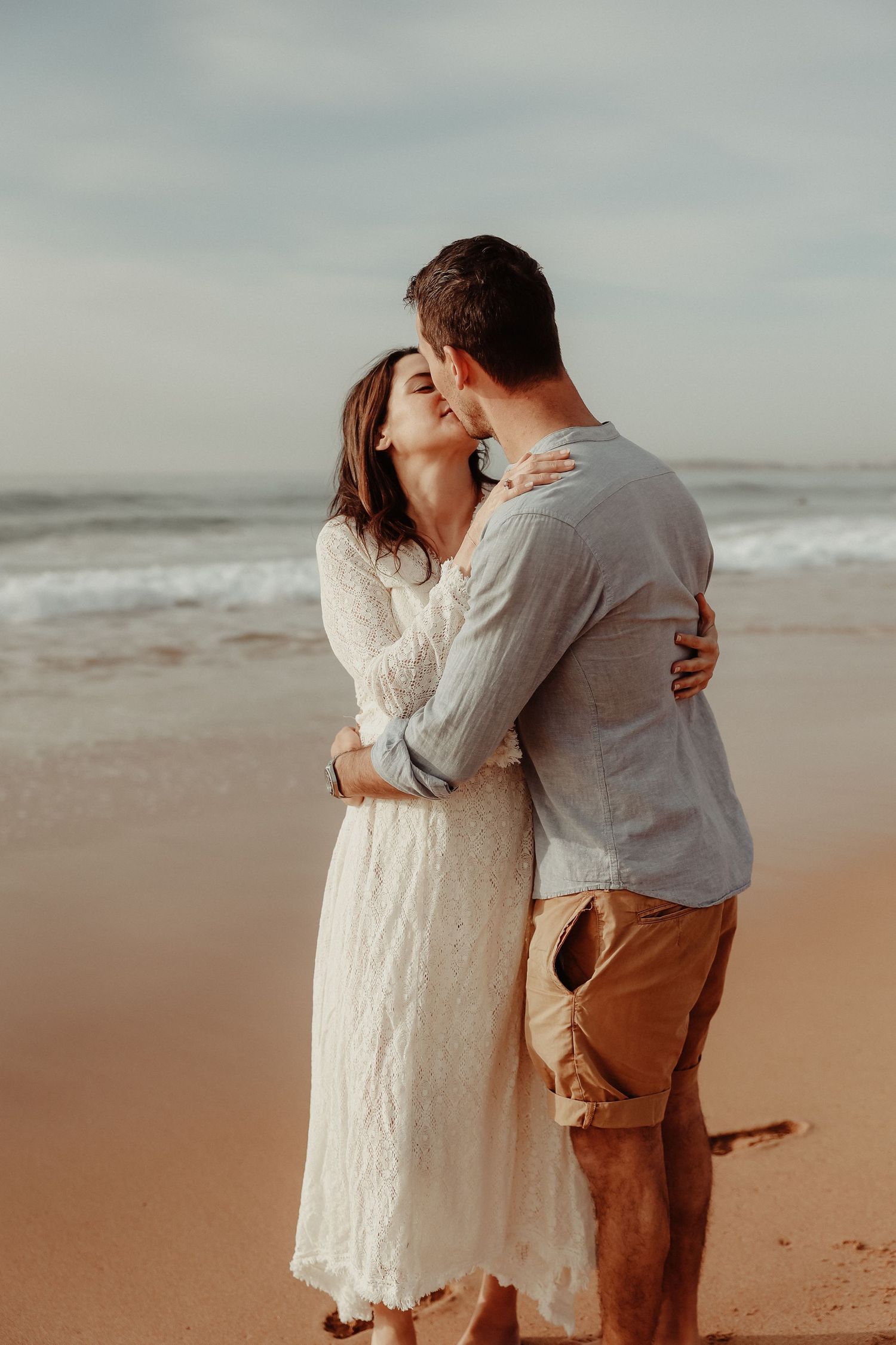 Couple sharing a romantic kiss on a sandy beach, woman in white lace dress embracing man in casual summer outfit.