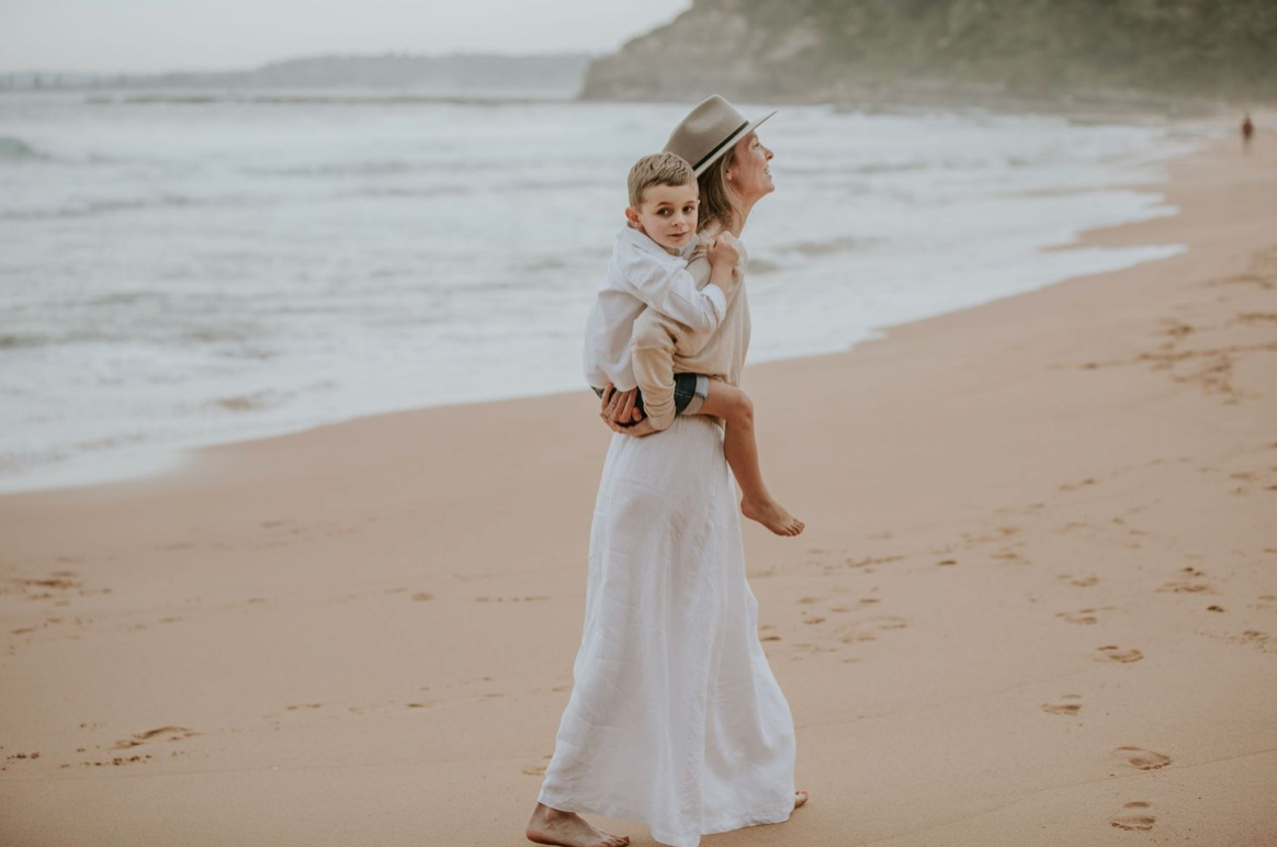 A woman in a white dress carries a baby while walking along a sandy beach.