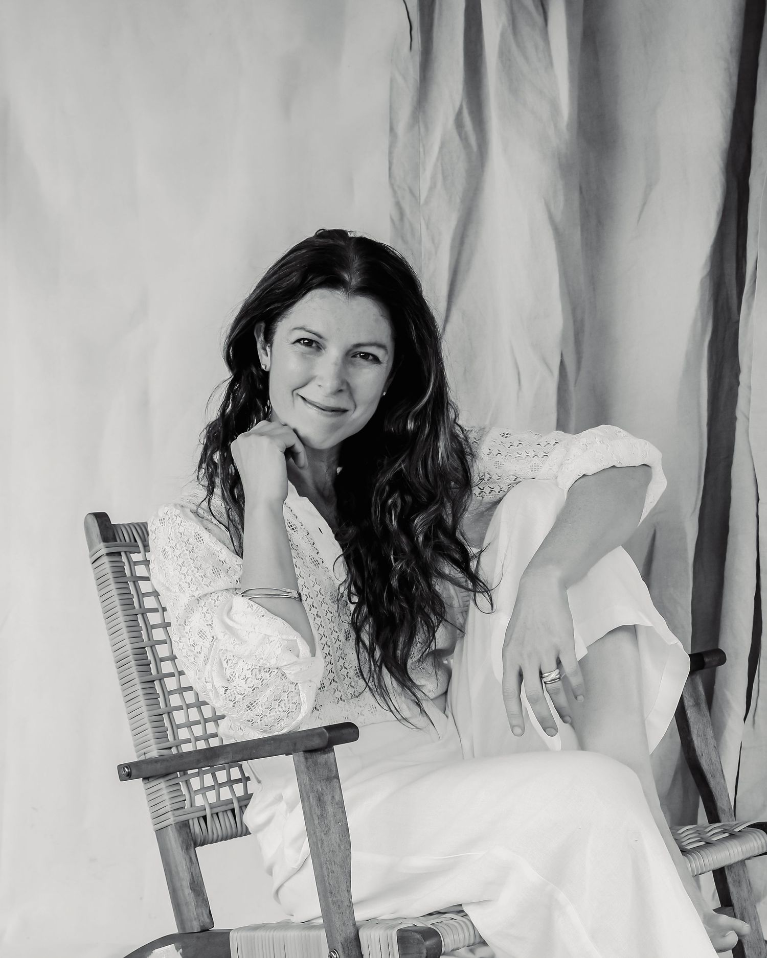 Black and white portrait of a smiling woman with long dark hair in white outfit seated in a wooden chair.