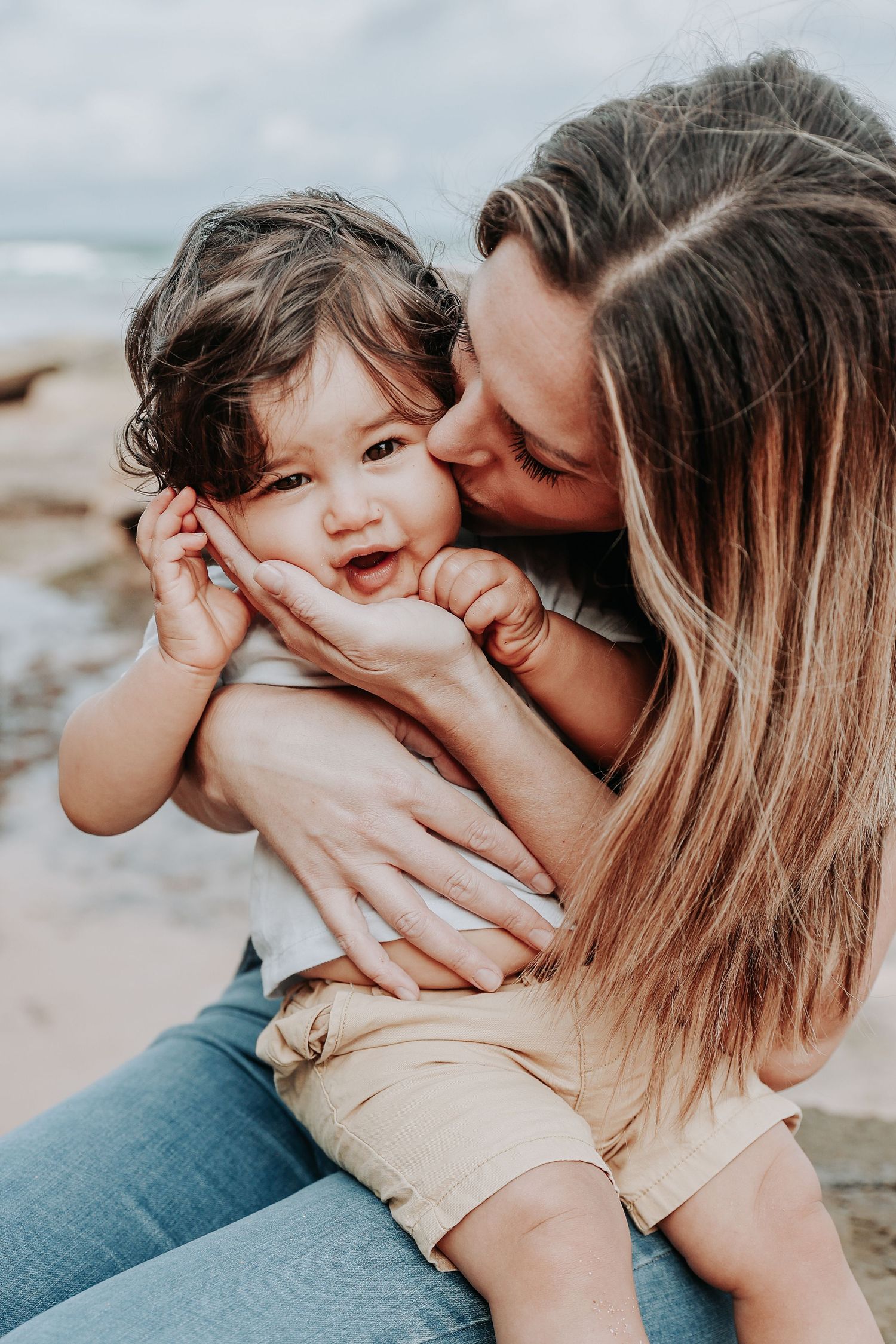 A loving mother kisses her happy toddler on the cheek at a rocky beach, holding the smiling child tenderly.