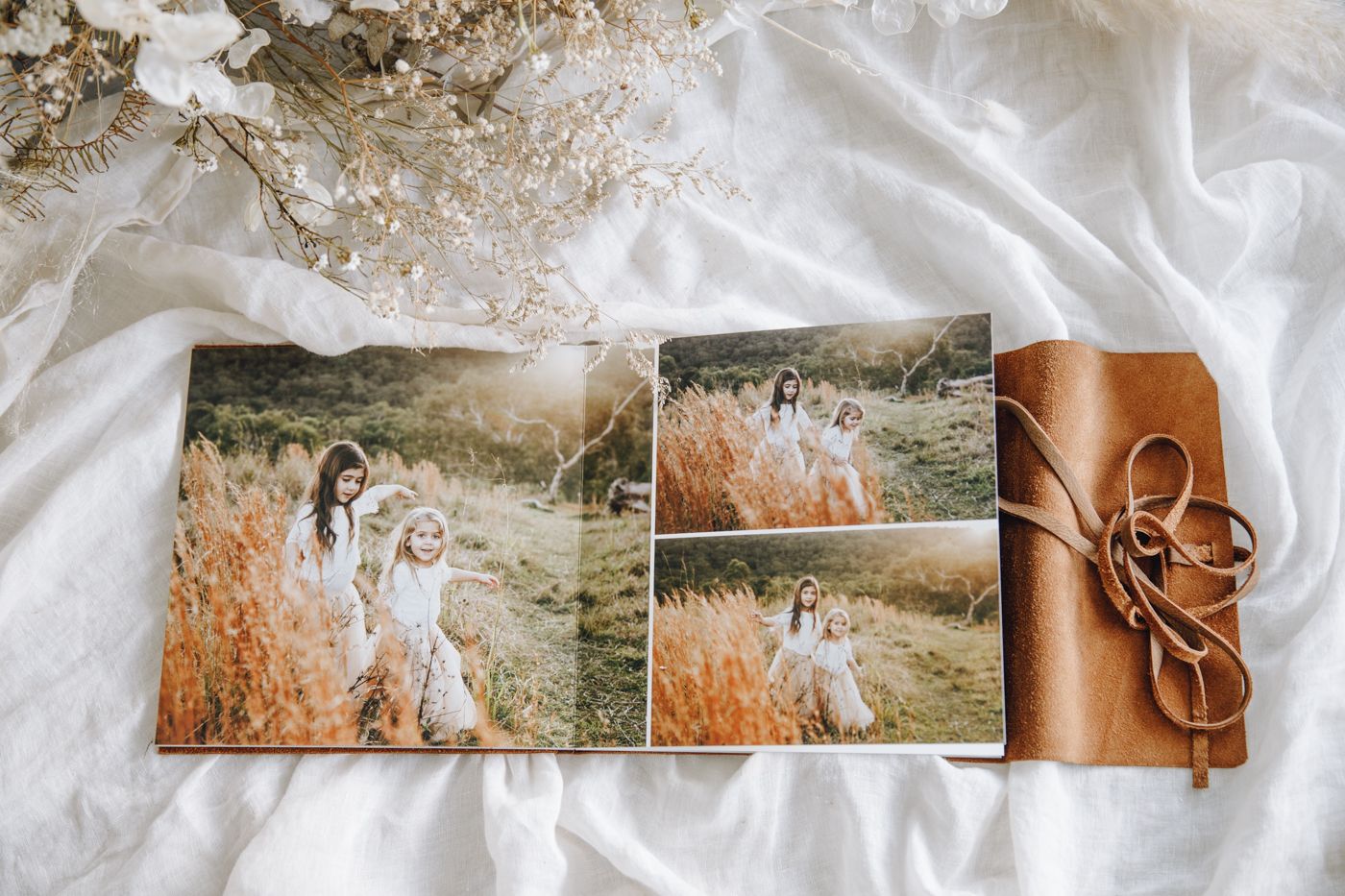 A leather-bound photo album open on white fabric displaying family portraits of a mother and daughter in golden fields.