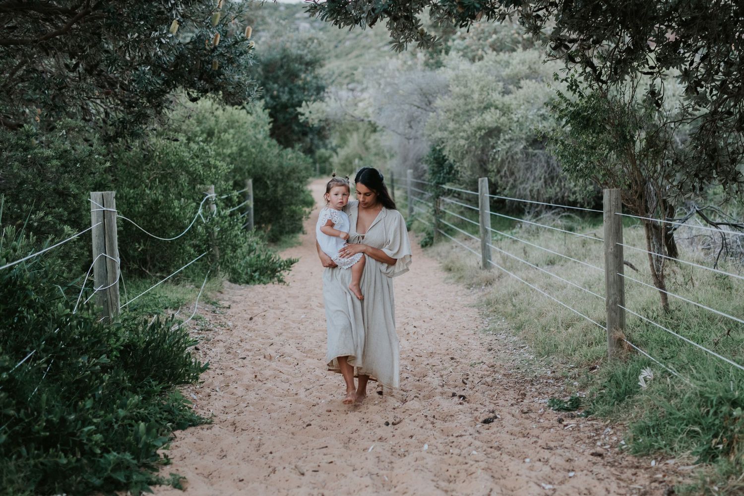 Mother in flowy dress holds toddler on scenic dirt path surrounded by lush greenery and wooden fence.