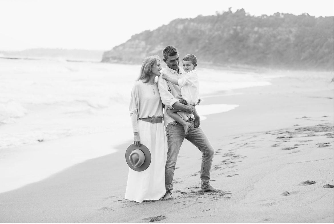 Black and white family photo of couple with toddler on a sandy beach with ocean waves.