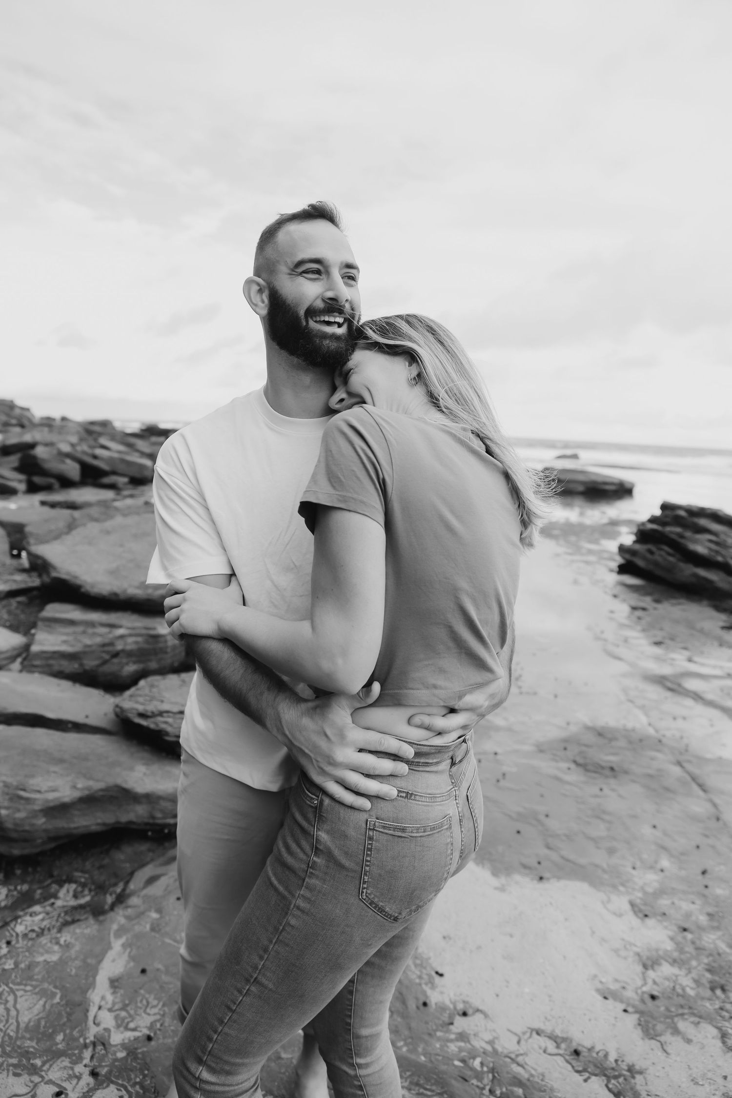 Black and white photo of a happy couple embracing on a rocky beach, the man smiling as the woman leans in close.