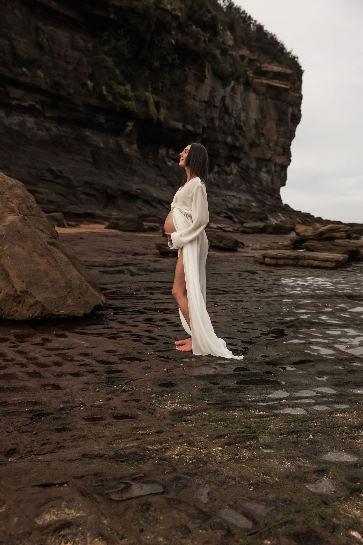 A pregnant woman in a flowing white gown poses on rocky coastal terrain near a cliff.