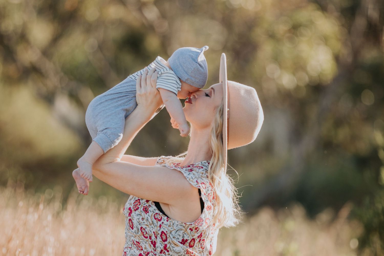 A blonde mother in a floral dress lifts her baby for a kiss in a sunny outdoor field.