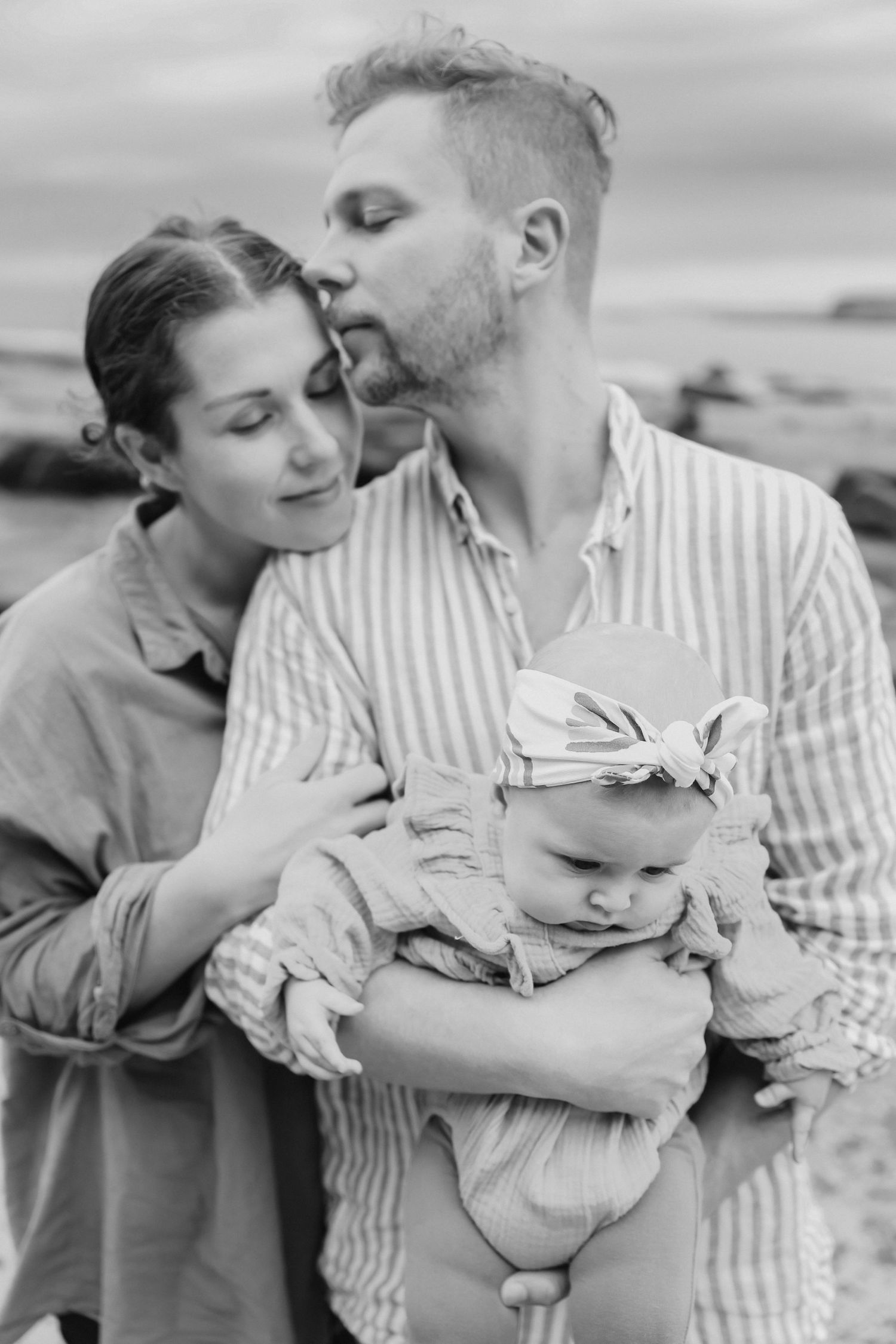 Black and white photo of a couple with their baby, man kissing woman's forehead outdoors near the ocean.