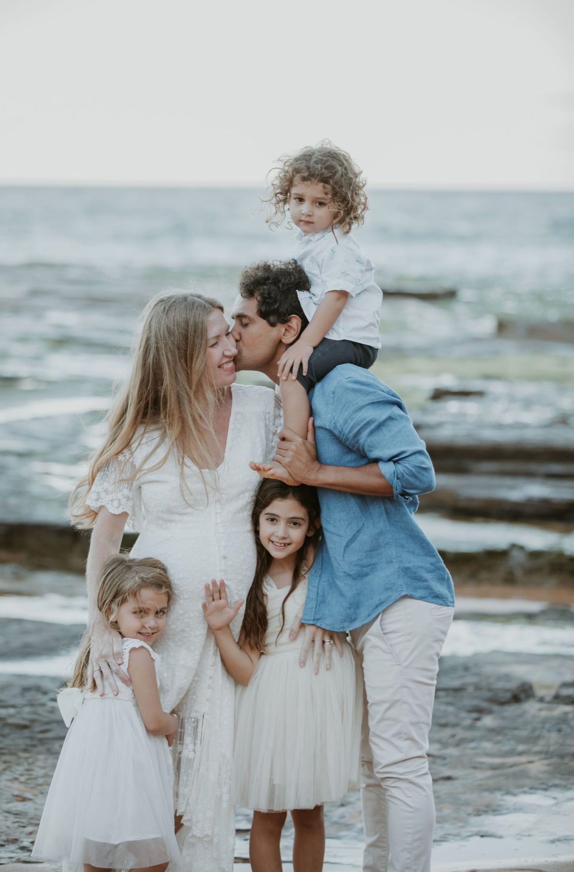 Family of five posing on a rocky beach, father kissing mother on the cheek.