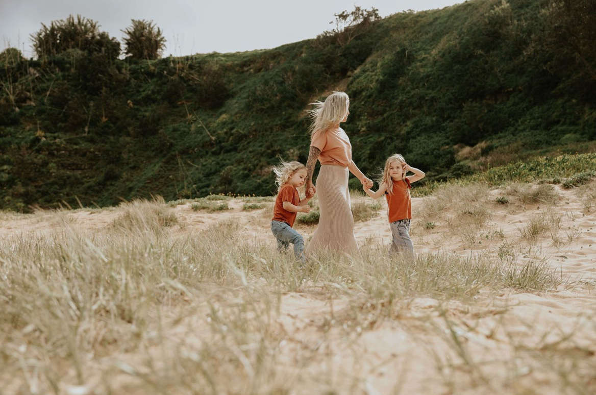 Mother walking with two young children through sandy coastal dunes holding hands together.