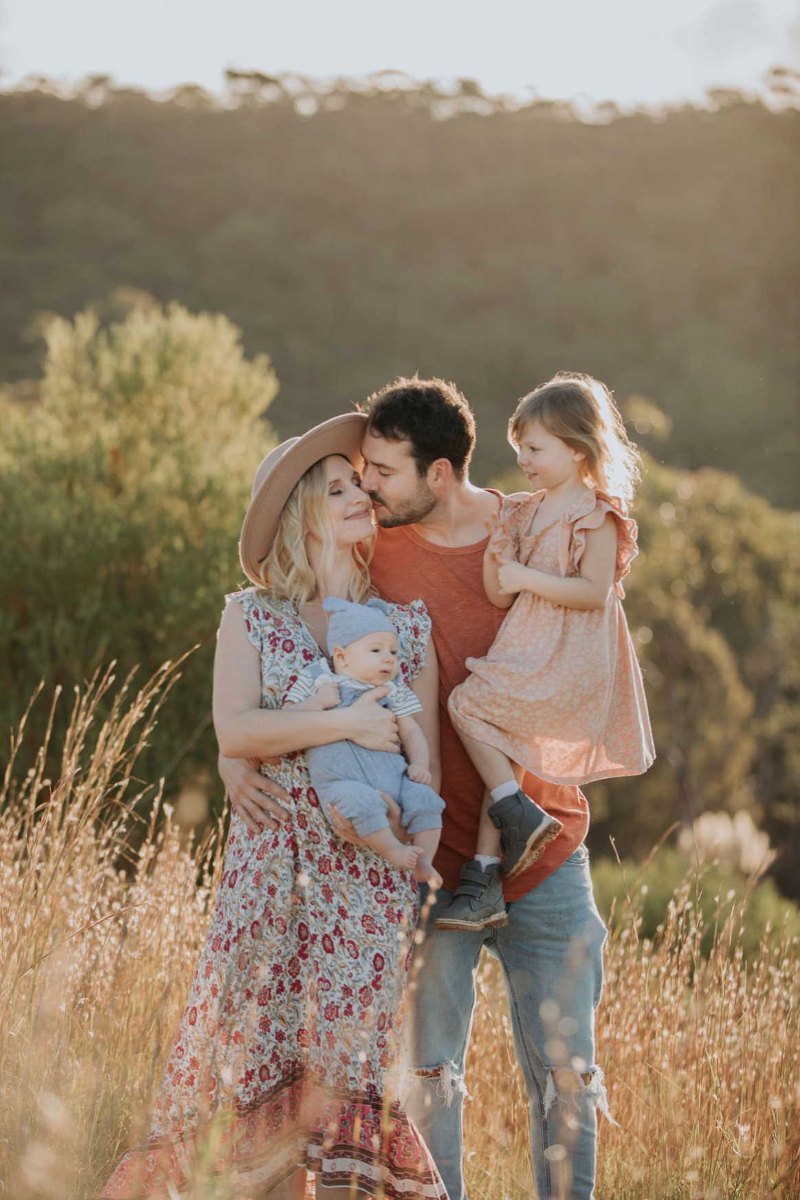 Family of four posing in golden field at sunset, parents holding two young children in bohemian style outfits.