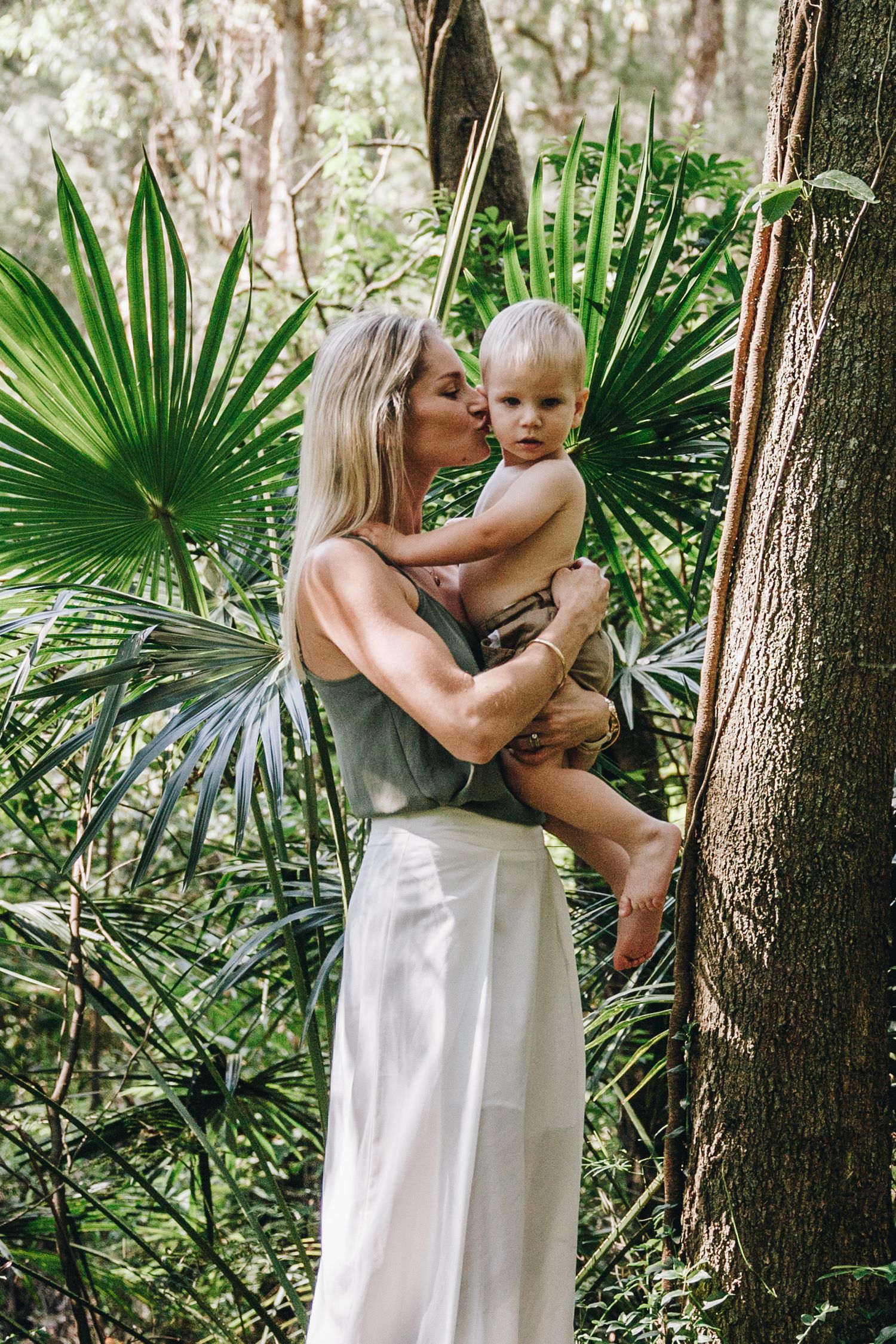 A blonde mother kisses her toddler son while holding him in a lush tropical forest setting.