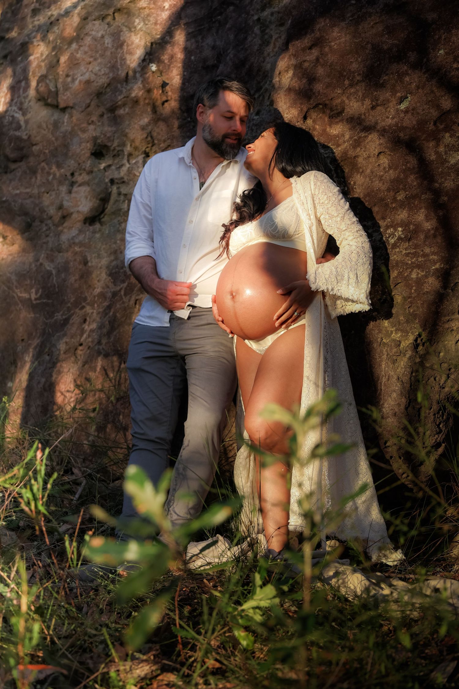 Couple leaning against rock face outdoors, man kissing pregnant woman's head in warm golden light.