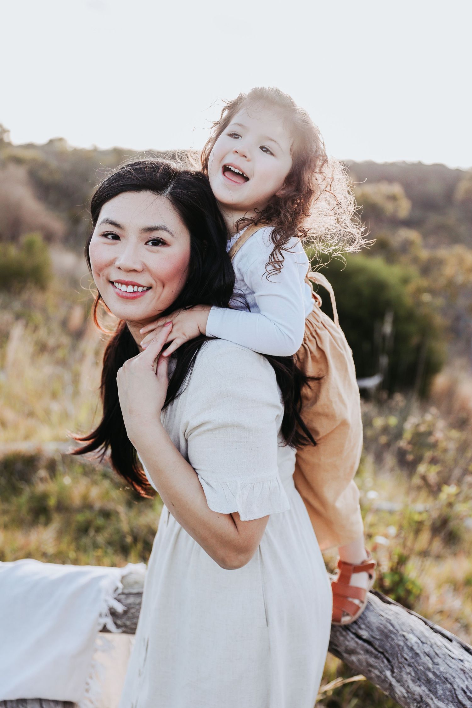 Smiling Asian mother in white dress with laughing curly-haired daughter on her back outdoors.