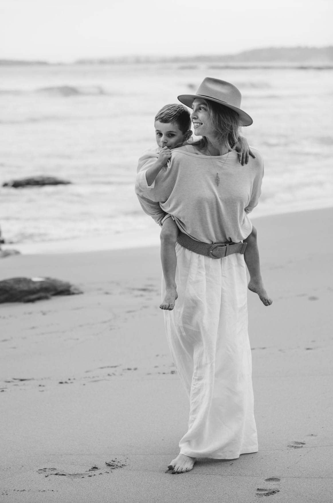 A woman in a wide-brim hat and white pants carries a child on her back while walking barefoot on a beach.