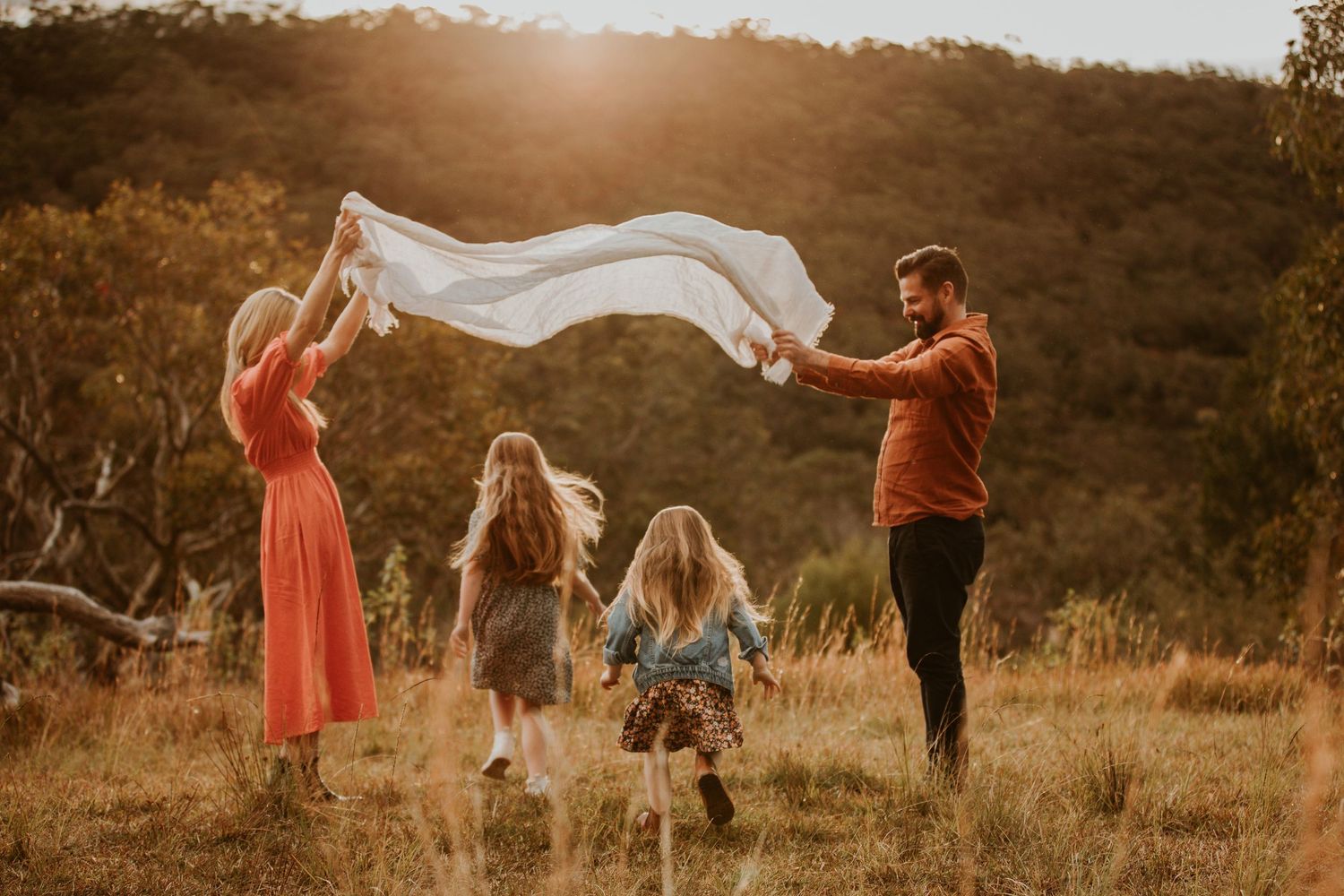 Family playing with white sheet in golden sunlit meadow with forest background.