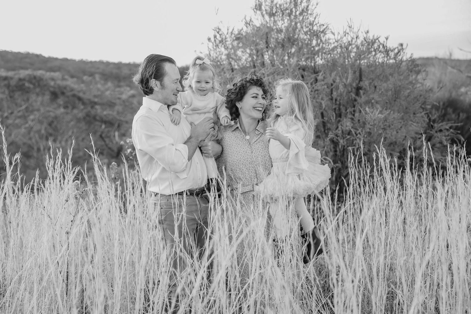 Black and white family photo of parents holding two young children in a field of tall grass with trees in background.