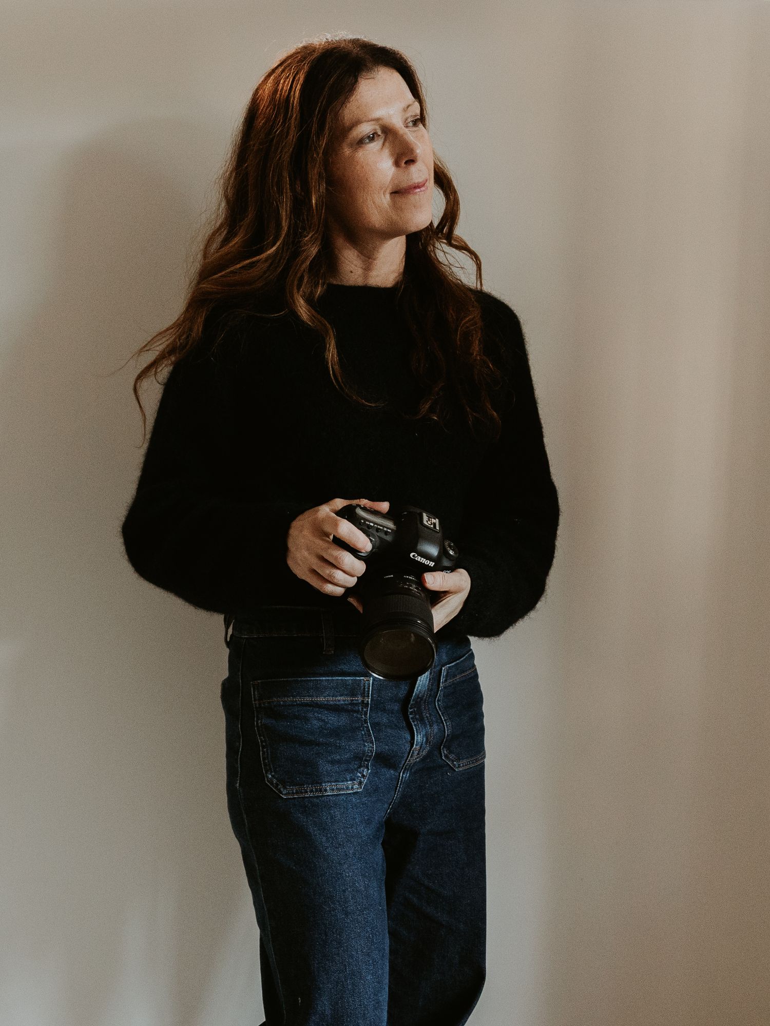 Woman with long brown hair holding a camera, wearing a black sweater and jeans, standing against a neutral background.