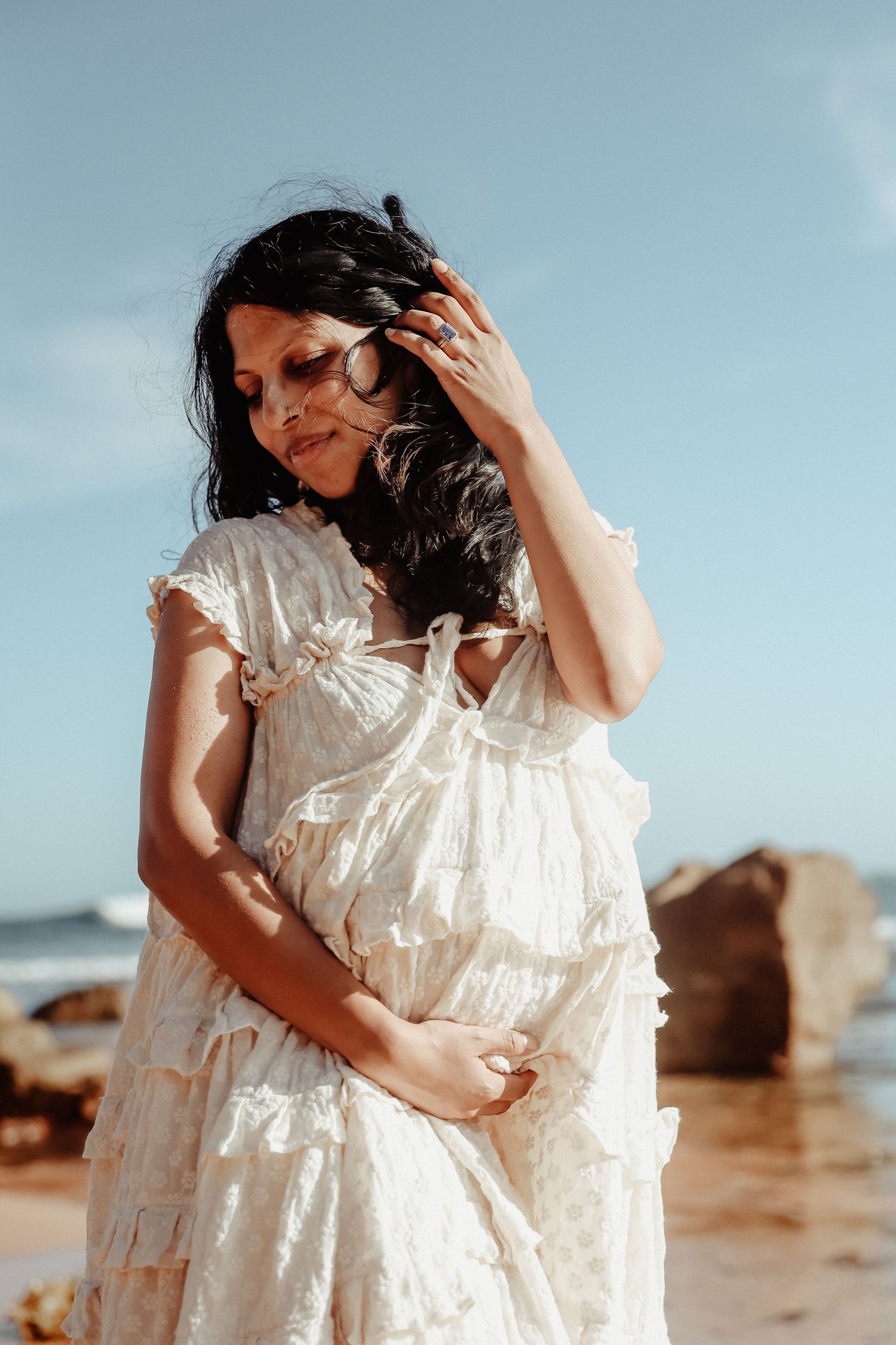 Pregnant woman in white ruffled dress touching her hair on a rocky beach under a clear blue sky.