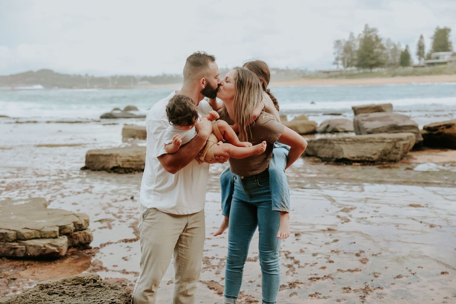 Young couple kissing on rocky beach while holding their two small children.