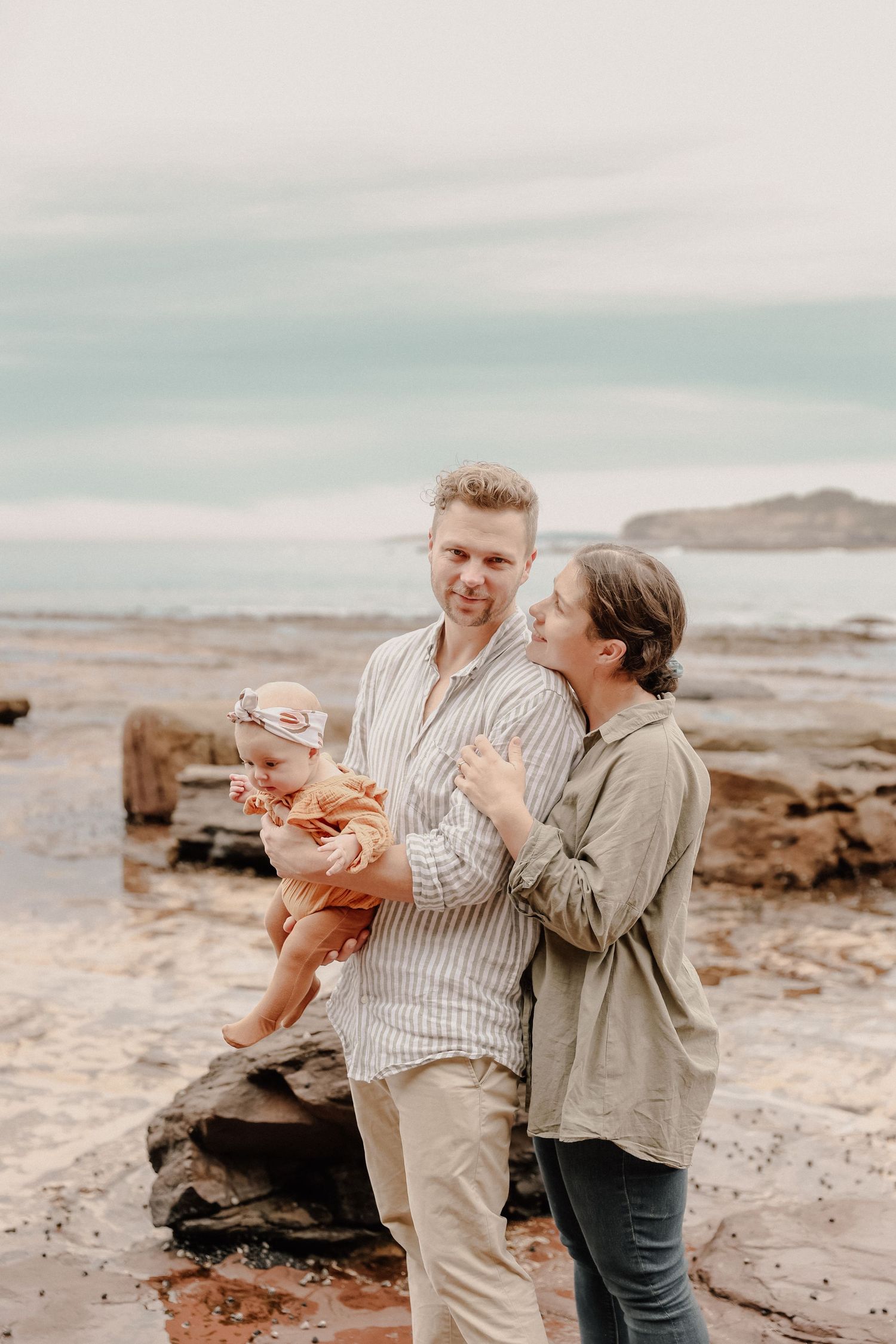 Young couple holding baby at rocky beach with ocean backdrop.