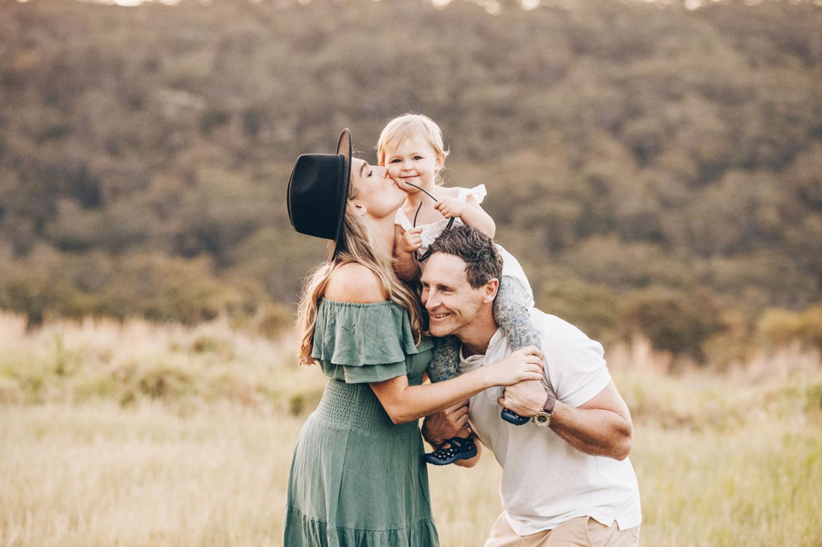Happy family of three in golden meadow, parents kissing baby on shoulders at sunset.