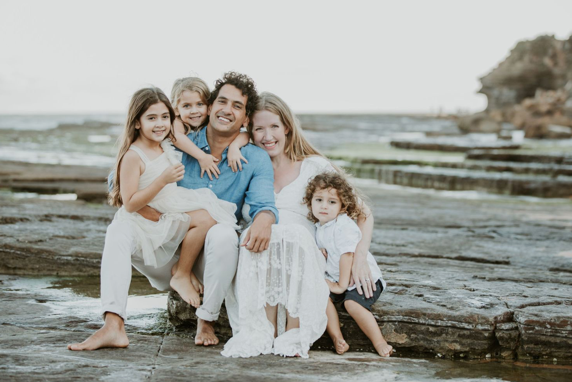 Happy family of five sitting together on rocky beach shoreline smiling.