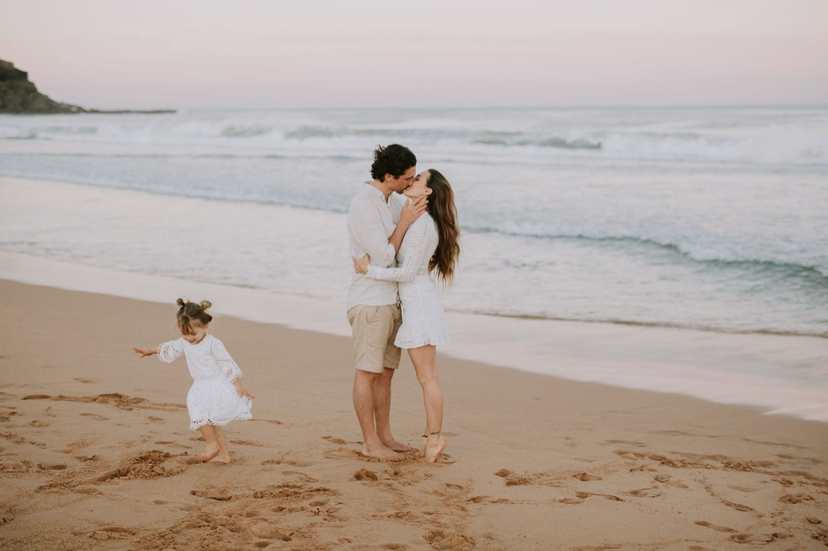 Couple sharing a kiss on a sandy beach at sunset while their toddler plays nearby in white dress.