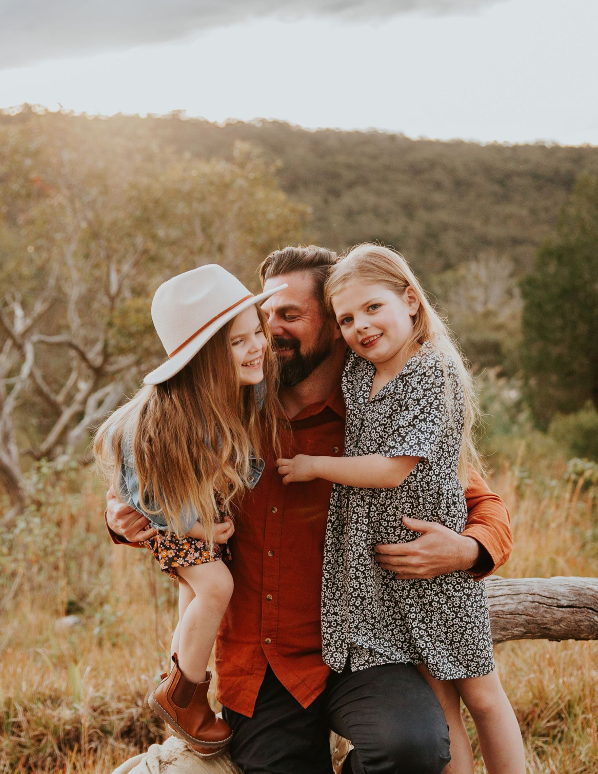 A bearded father holds two smiling daughters in a sunlit outdoor nature setting.