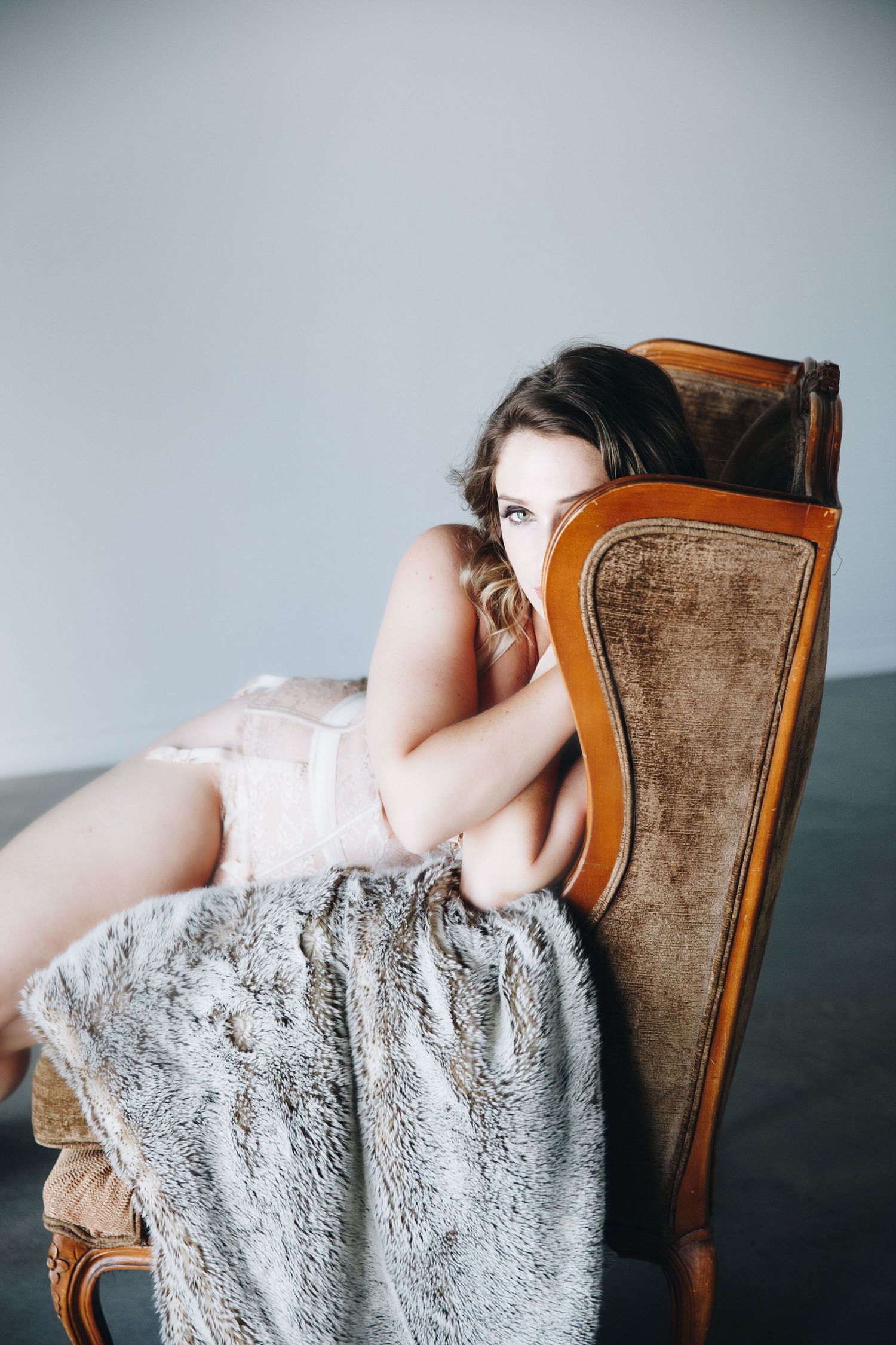 Young woman in blush lingerie reclining on vintage velvet armchair with fur blanket.
