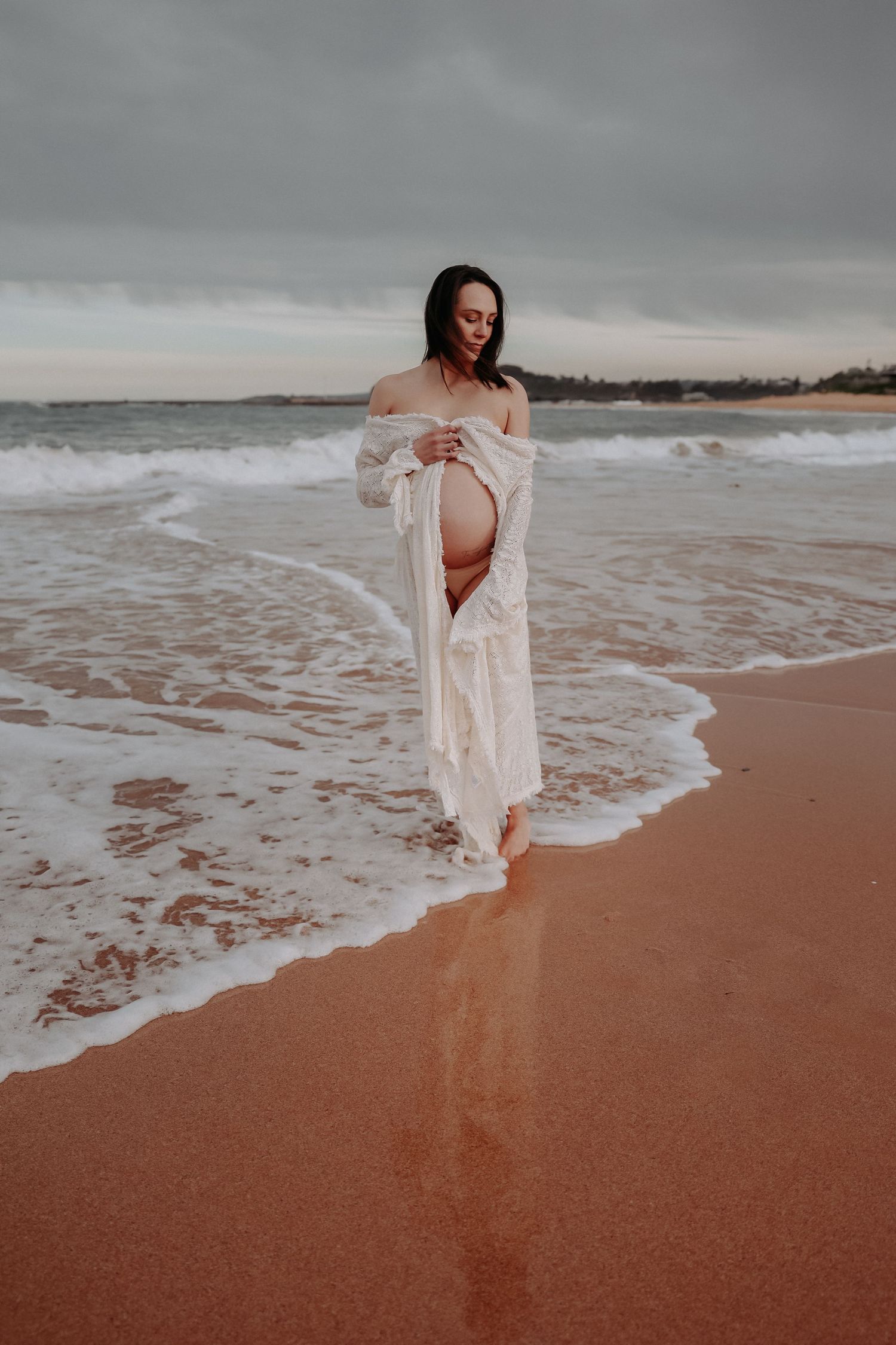 Pregnant woman in white robe standing on sandy beach with ocean waves, showcasing her baby bump.