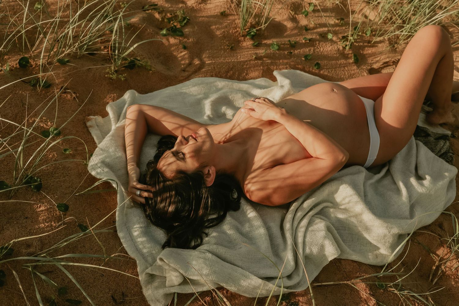 Pregnant woman relaxing on a blanket outdoors on sandy ground surrounded by grass in warm sunlight.