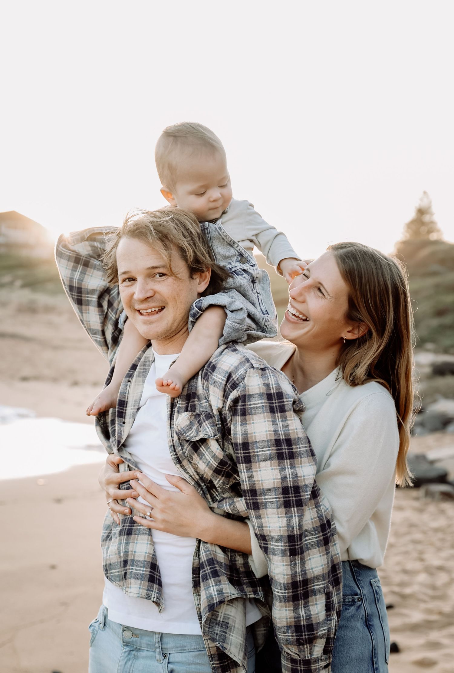 Young couple laughing on beach with baby on shoulders at golden hour.