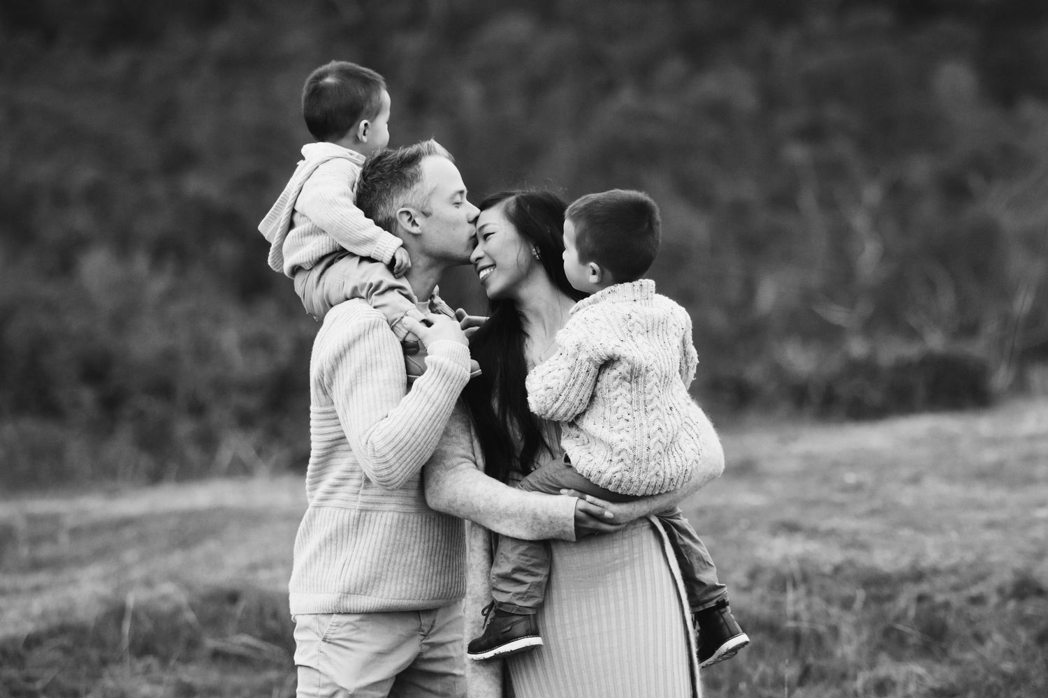 Black and white family photo of parents kissing while holding two young children in an outdoor field setting.
