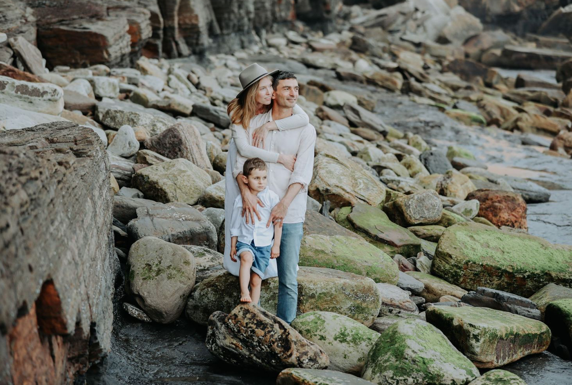 Family of three standing on rocky coastal shoreline for a portrait.