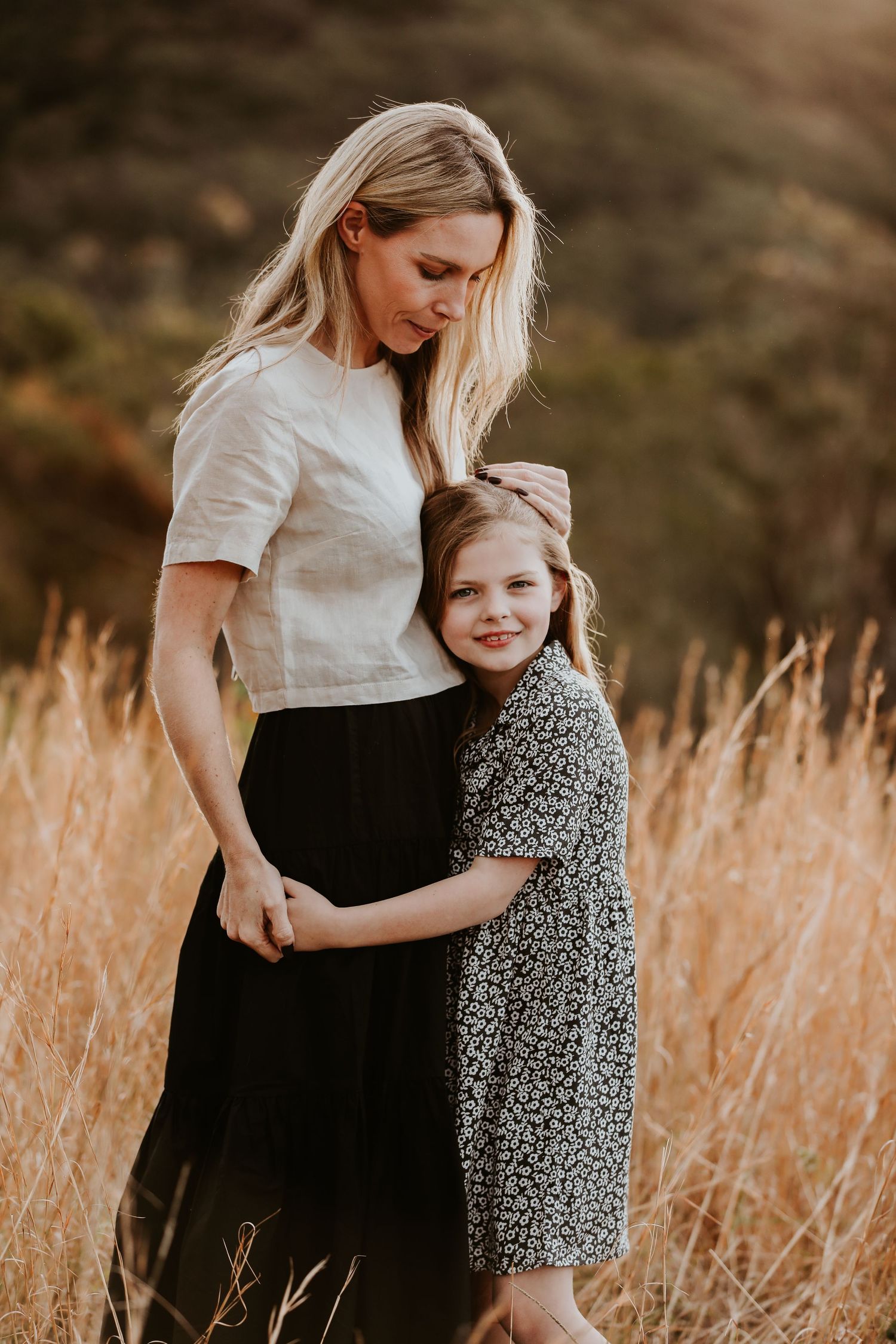Blonde mother in white top and black skirt embraces smiling young daughter in floral dress outdoors.