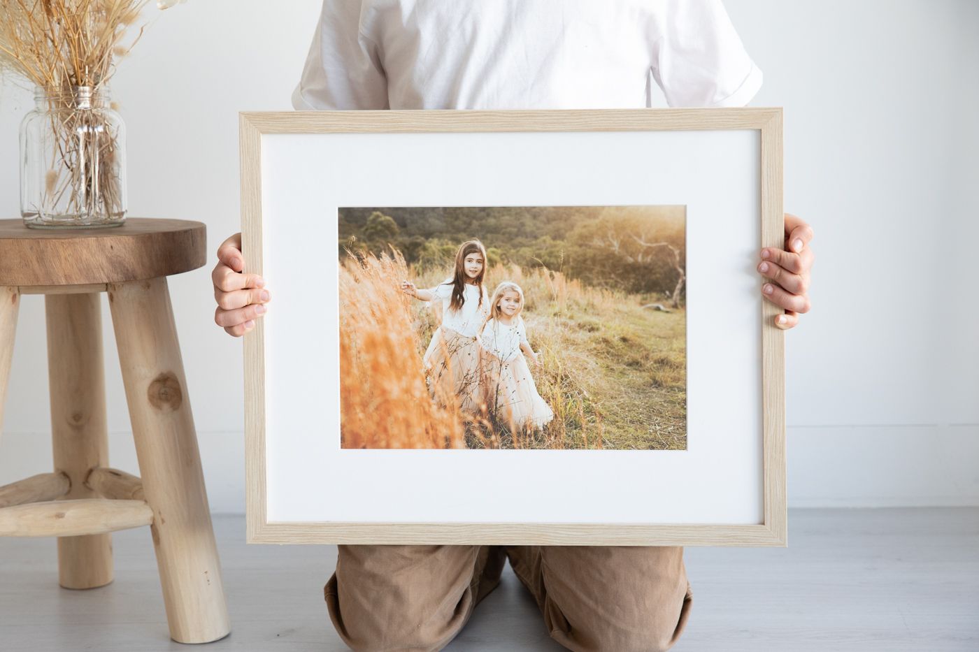 Person holding a framed photo of two girls in a golden field, displayed with a white mat in a natural wood frame.