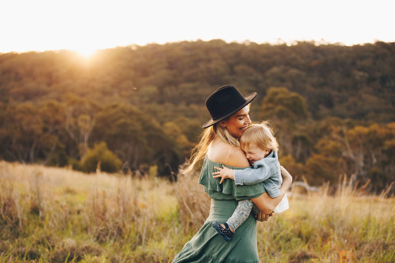A mother in a green dress and black hat joyfully holds her toddler in a sunlit field with trees behind.