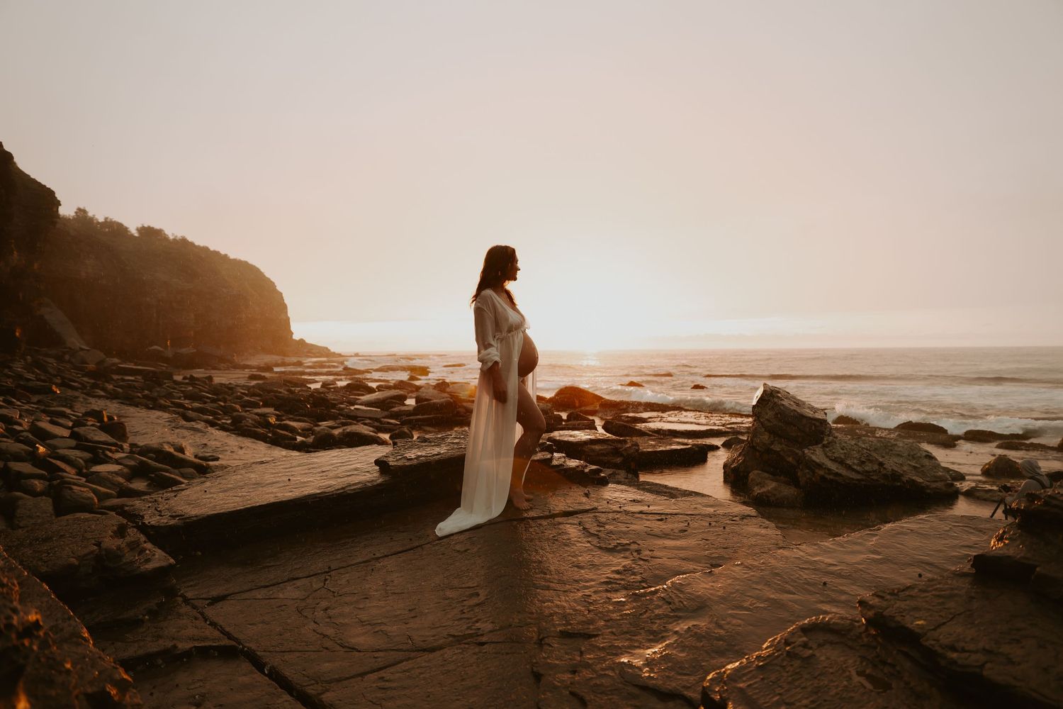 Pregnant woman in white gown standing on rocky beach at golden sunset by the ocean.