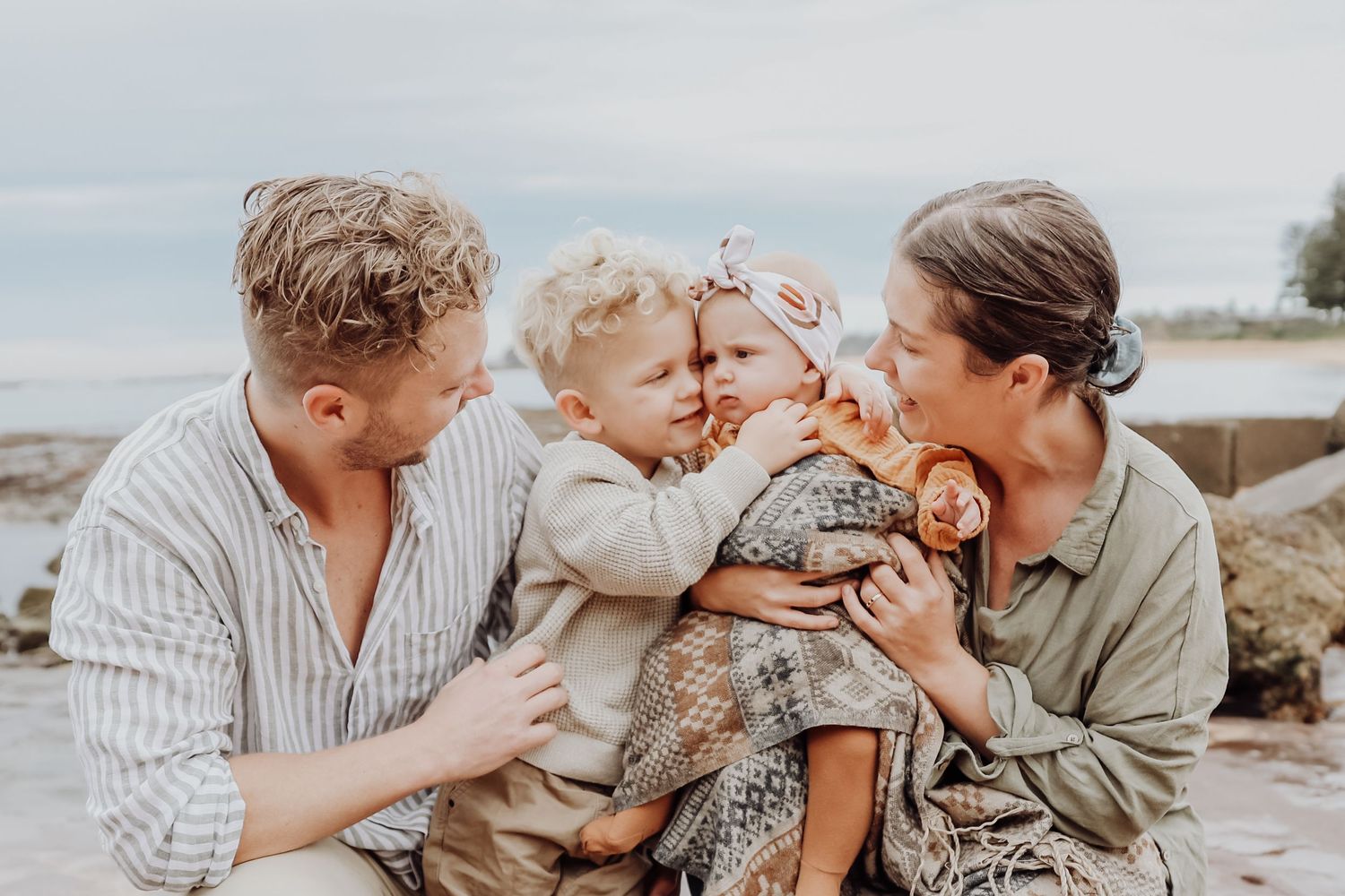 Family of four enjoying a beach day, parents smiling as toddler hugs baby girl in floral wrap.