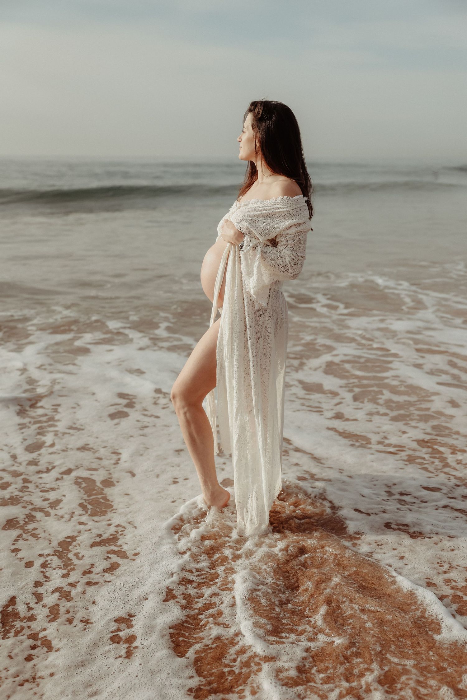 Pregnant woman in white lace wrap standing in ocean waves on sandy beach.